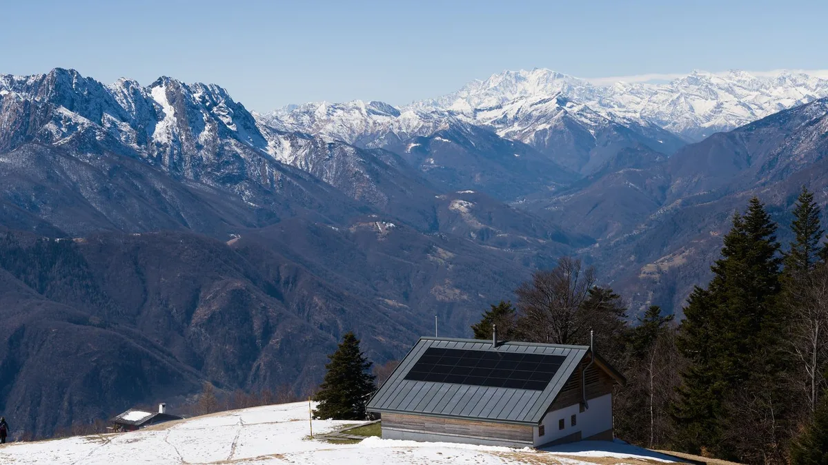 Cabană la munte cu panouri fotovoltaice pe acoperiș, pe o pajiște cu zăpadă, cu creste înzăpezite în fundal