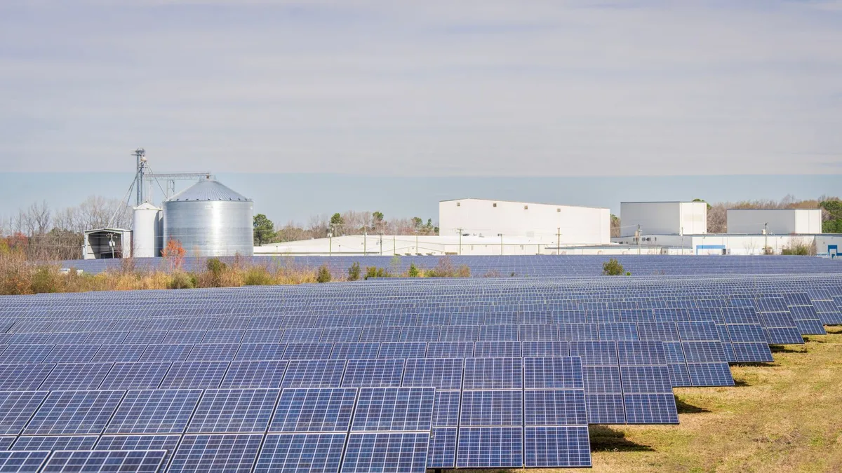 Panouri fotovoltaice montate pe sol lângă silozuri de cereale și hale agricole